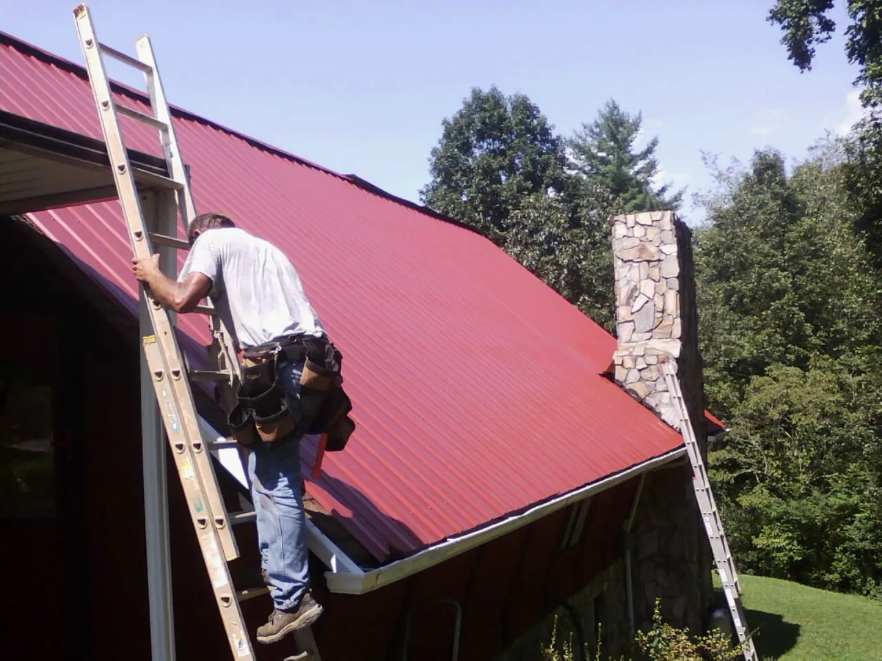 Roofer on a ladder beside a red metal roof and stone chimney — Soffit & Fascia Repair by Roofing Brothers in Greeneville